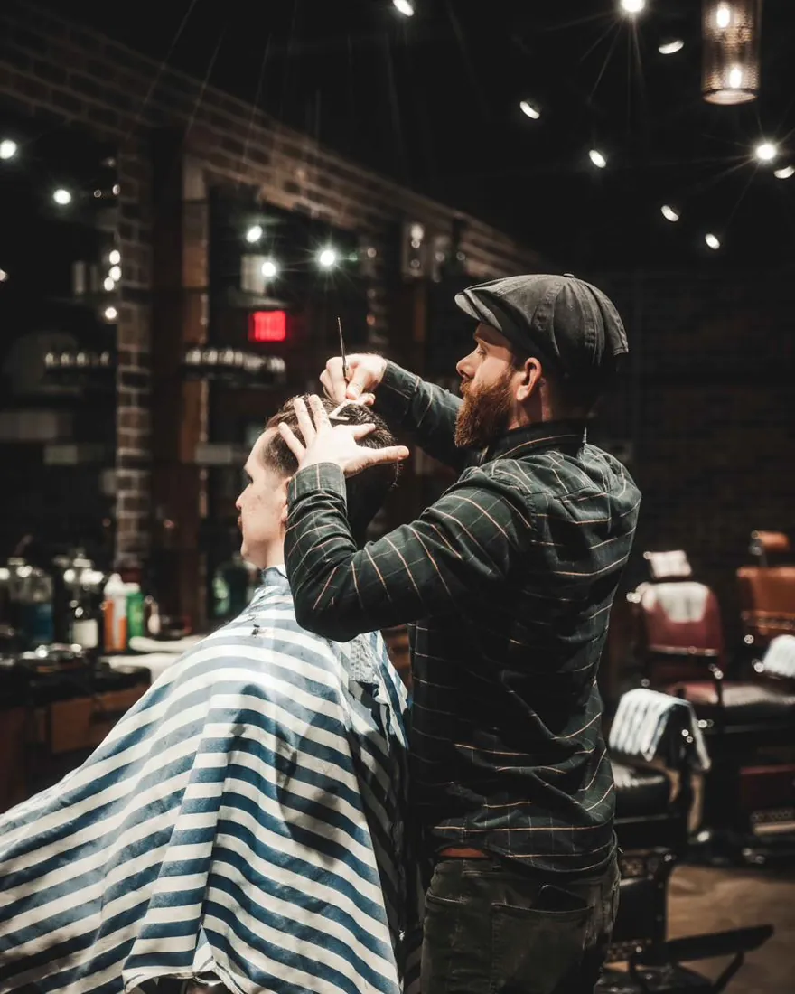 A barber in a flat cap cutting a client’s hair with scissors