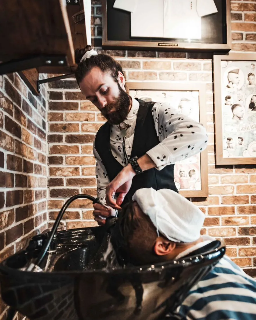 A bearded barber washing a client’s hair at the basin