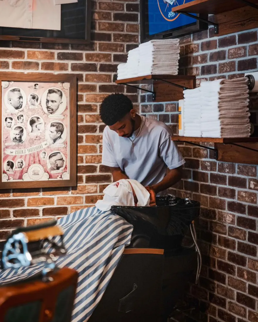 A barber rinsing a client’s hair at the basin beside vintage haircut posters