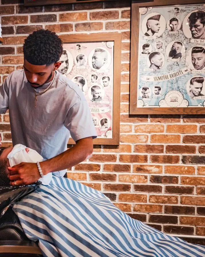 A barber at the wash basin beside stacked towels and vintage haircut posters