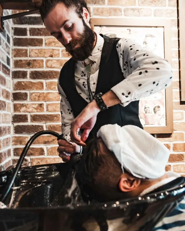 A bearded barber washing a client’s hair at the basin